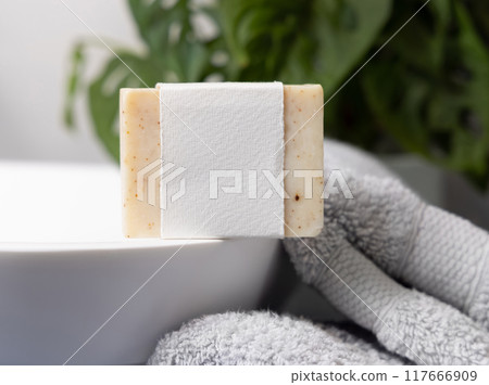 Soap bar with blank label on vessel sink near grey towels and green plant close up, mockup 117666909