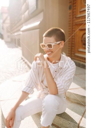 Hipster fashion woman in stylish glasses sits on a curb in the street in Europe. 117667497