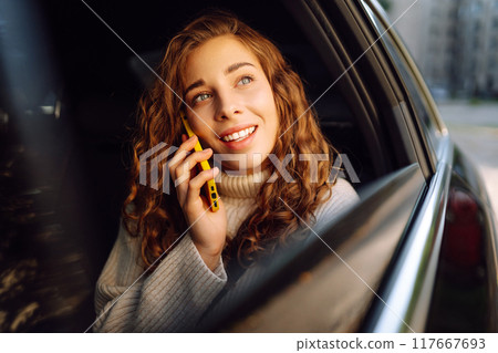 Positive young woman in casual clothes sits in the back seat of a car with a mobile phone. Positive young woman in casual clothes sits in the back seat of a car with a mobile phone. 117667693