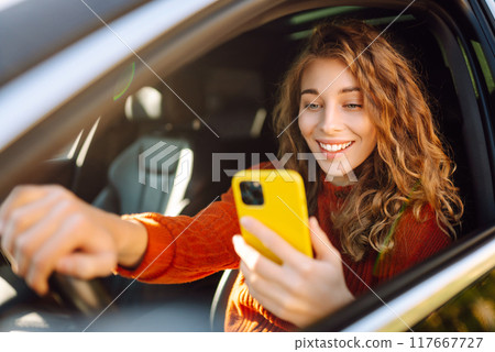Portrait of a young woman sitting in a car in the driver's seat looking into a smartphone. Portrait of a young woman sitting in a car in the driver's seat looking into a smartphone. 117667727