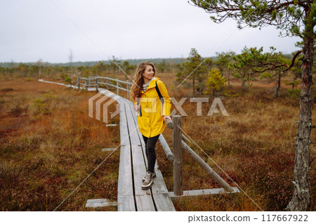 Smiling female traveler in yellow coat walks along wooden path among wild nature. Travel, vacation. 117667922
