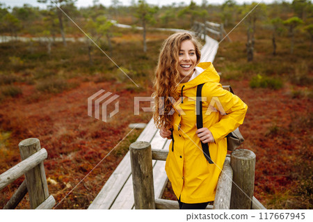 Smiling female traveler in yellow coat walks along wooden path among wild nature. Travel, vacation. Smiling female traveler in yellow coat walks along wooden path among wild nature. Travel, vacation. 117667945
