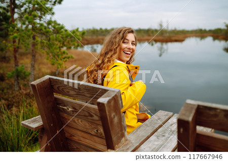 Smiling female traveler in yellow coat walks along wooden path among wild nature. Travel, vacation. 117667946