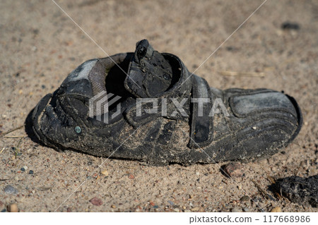 An old, dirty, wrinkled, black shoe, lying on a gravel road 117668986