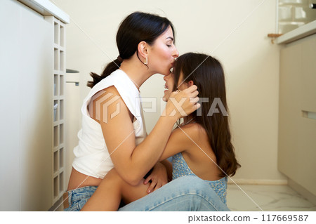 A woman shares a tender moment with a girl sitting on the kitchen floor in a cozy, well-lit home A woman shares a tender moment with a girl sitting on the kitchen floor in a cozy, well-lit home 117669587