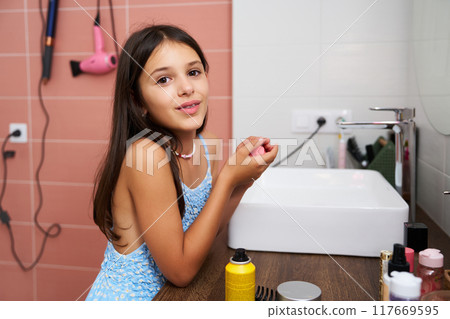 Young girl applying lip balm in a bathroom with a sink and hair dryer 117669595