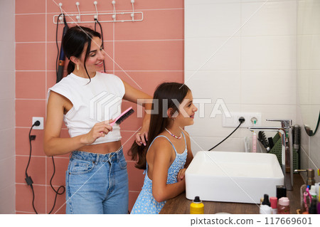 Mother brushes daughter's hair in a modern bathroom during a sunny afternoon 117669601