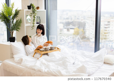 Mother and daughter having breakfast in bed by window 117670340