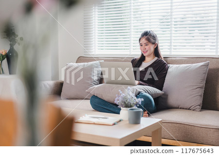 Smiling young Asian woman uses laptop while sitting on couch, enjoying comfortable and relaxed moment at home. 117675643