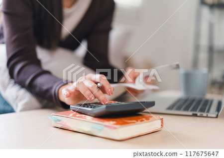 Close up of woman using calculator and holding receipts while managing finances at home. Close up of woman using calculator and holding receipts while managing finances at home. 117675647