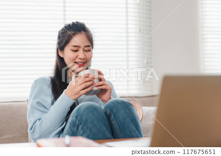 Asian woman sitting comfortably with a cup of hot beverage, smiling and enjoying a cozy moment in living room. 117675651