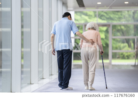 Rear view of a senior woman walking with a cane and a caregiver 117676160
