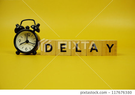 Delay with wooden blocks alphabet letters and alarm clock on yellow background Delay with wooden blocks alphabet letters and alarm clock on yellow background 117676380