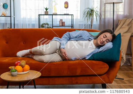 Smiling young pregnant woman stroking big belly while sleeping on comfortable sofa dreaming at home 117680543