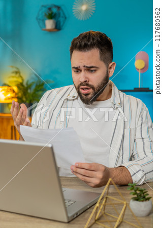 Amazed businessman opening envelope reading bad news in letter feels upset bankruptcy at home office 117680562
