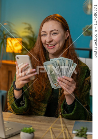 Smiling happy woman counting money dollar cash use smartphone income saves lottery win at home Smiling happy woman counting money dollar cash use smartphone income saves lottery win at home 117680571