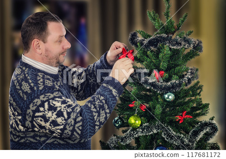 The Art of Christmas Decorating: Gentleman in Patterned Jumper Demonstrates Care and Tradition in Tree Trimming. The Art of Christmas Decorating: Gentleman in Patterned Jumper Demonstrates Care and Tradition in Tree Trimming. 117681172