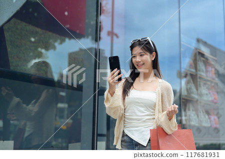 Smiling young asian woman with shopping bags using mobile phone on the city street 117681931