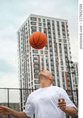Sporty adult man playing with orange basketball ball on sports court at courtyard. Street and urban style. Focus on ball. Fun exercise. Sporty adult man playing with orange basketball ball on sports court at courtyard. Street and urban style. Focus on ball. Fun exercise. 117682584