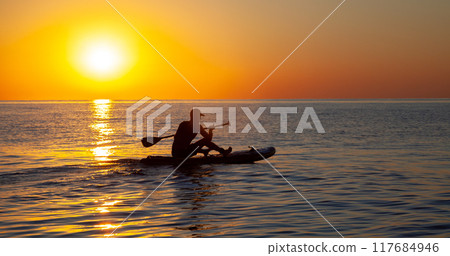 Silhouette of a man floating on a board with an oar. Dawn sun on the sea. Swimming on a supboard. Water active sports. Beautiful landscape. Sunset on the sea. Ocean coast. Copy space. 117684946