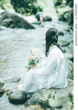 Back view of a woman in a white dress sitting by a river in summer 117685060