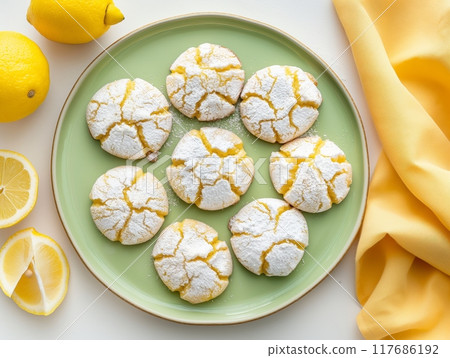 A green plate holds eight lemon crinkle cookies dusted with powdered sugar. Fresh lemons and lemon slices are placed nearby, with a yellow cloth draped to the side. 117686192