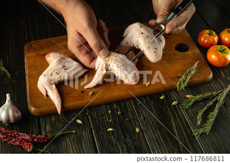 Chef prepares delicious chicken wings on the kitchen table with spices. Fork in the hands of the cook. Concept of cooking Buffalo wings for lunch in a restaurant or hotel Chef prepares delicious chicken wings on the kitchen table with spices. Fork in the hands of the cook. Concept of cooking Buffalo wings for lunch in a restaurant or hotel 117686811