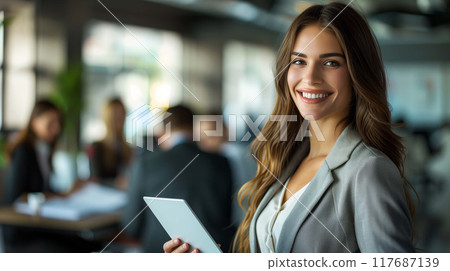 Businesswoman stands confidently with a smile and a tablet, while her colleagues sit in a meeting 117687139
