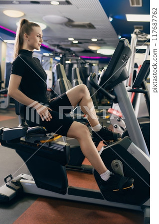 Young woman exercising on a stationary bike in a modern gym during daytime Young woman exercising on a stationary bike in a modern gym during daytime 117687762
