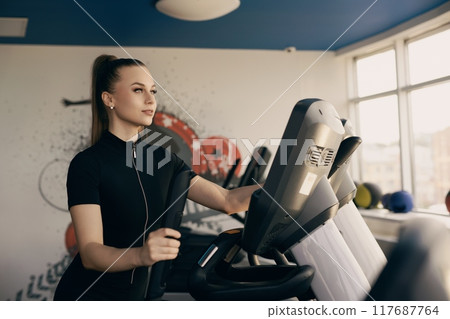 Young woman exercising on elliptical machine in modern gym during daytime Young woman exercising on elliptical machine in modern gym during daytime 117687764