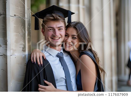 A young man wearing a black graduation gown and cap is posing with his girlfriend stand in front of a university's corridor, showing love for each other, look happy after just having graduated A young man wearing a black graduation gown and cap is posing with his girlfriend stand in front of a university's corridor, showing love for each other, look happy after just having graduated 117687909
