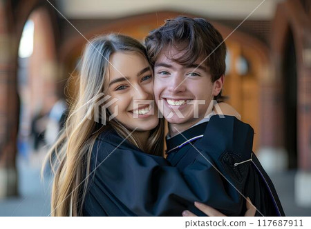 A young man wearing a black graduation gown and cap is posing with his girlfriend stand in front of a university's corridor, showing love for each other, look happy after just having graduated A young man wearing a black graduation gown and cap is posing with his girlfriend stand in front of a university's corridor, showing love for each other, look happy after just having graduated 117687911