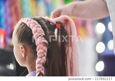 The master braids the child's pigtails from a pink kanekalon. Close-up, side view. The hands of the master weave the kanekalon material into the hair. High quality photo The master braids the child's pigtails from a pink kanekalon. Close-up, side view. The hands of the master weave the kanekalon material into the hair. High quality photo 117692217