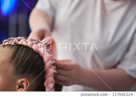 Close-up of the pigtail on the child's head made of pink kanekalon. The hands of the master who weave the kanekalon material into the hair. With space to copy. High quality photo 117692224