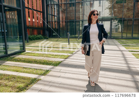 A stylish businesswoman confidently walks outdoors in a modern office complex during the day 117696678