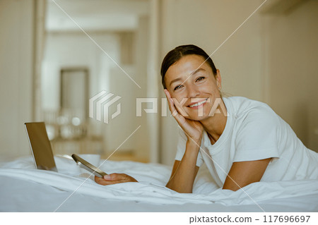 A young woman relaxes in her cozy bed, engaged with a laptop and phone, enjoying leisure A young woman relaxes in her cozy bed, engaged with a laptop and phone, enjoying leisure 117696697