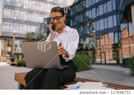A businesswoman works outdoors on her laptop while on the phone, balancing tasks effectively A businesswoman works outdoors on her laptop while on the phone, balancing tasks effectively 117696731