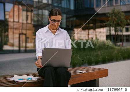 A modern businesswoman is efficiently working outdoors on her laptop while enjoying nature 117696746