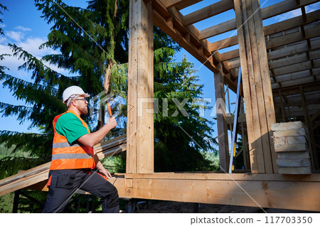 Carpenter constructing wooden frame house near the forest. Bearded man hammering nails into structure while wearing protective helmet and construction vest. Concept of modern ecological construction. 117703350