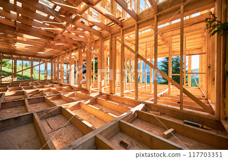 Under-construction residential wooden frame home located near a forest. Commencement of a new construction project for a cozy house or a mountain cottage. Idea of contemporary ecological construction. 117703351