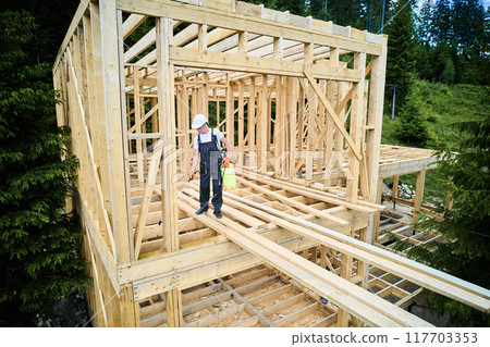 Laborer constructing wooden frame house near forest. Man treating woods, applying fire retardant using sprayer, while dressed in protective suit, helmet. Concept of modern eco-friendly construction. Laborer constructing wooden frame house near forest. Man treating woods, applying fire retardant using sprayer, while dressed in protective suit, helmet. Concept of modern eco-friendly construction. 117703353