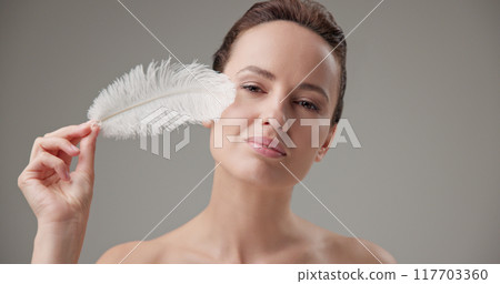 Beauty, health, cosmetics, anti-aging therapy and skin care concept - young beautiful brunette Caucasian woman holding a white fluffy feather, concept of tenderness, softness and lightness 117703360
