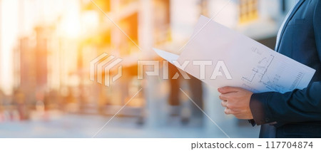 Business professional reviewing documents at a construction site during sunset, symbolizing growth and development. Business professional reviewing documents at a construction site during sunset, symbolizing growth and development. 117704874