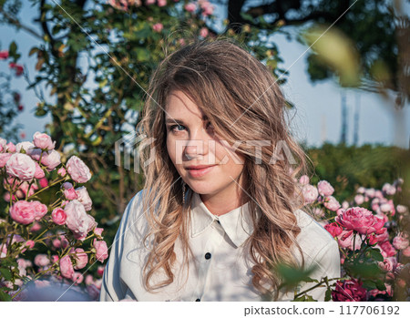 Young woman surrounded by blooming pink roses enjoying nature in a beautiful garden 117706192