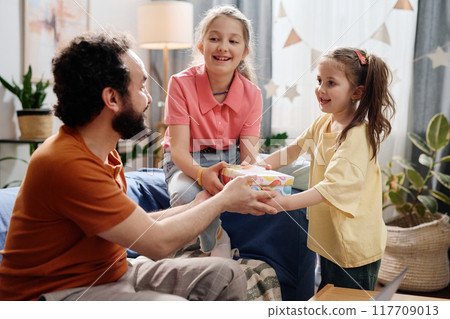 Father Giving Gift to Two Happy Young Girls at Home Father Giving Gift to Two Happy Young Girls at Home 117709013