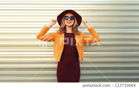 Beautiful smiling young woman wearing orange jacket, round hat on gray background 117710064