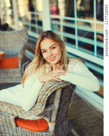 Portrait of beautiful blonde smiling young woman sitting on chair waiting in city cafe Portrait of beautiful blonde smiling young woman sitting on chair waiting in city cafe 117710095