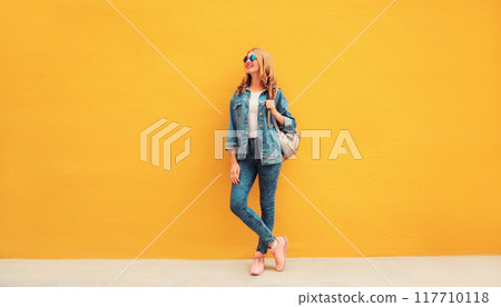 Full length beautiful smiling young woman wearing denim jacket, backpack on yellow background Full length beautiful smiling young woman wearing denim jacket, backpack on yellow background 117710118