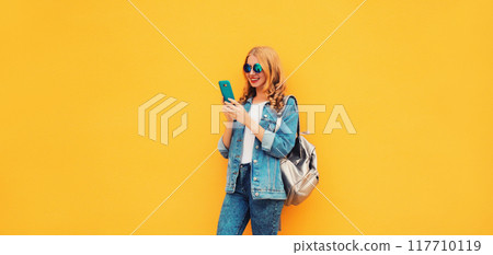 Portrait of stylish modern smiling young woman with smartphone in jean jacket on yellow background Portrait of stylish modern smiling young woman with smartphone in jean jacket on yellow background 117710119