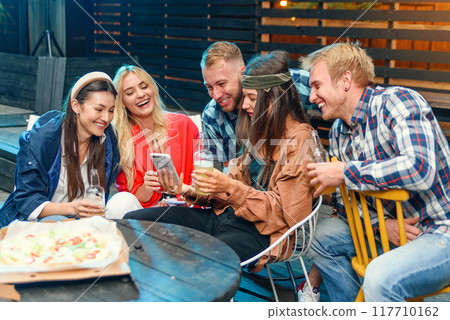 Beautiful woman shows interesting stuff on her smartphone to her friends while they spend time together on a weekend party outdoors. Beautiful woman shows interesting stuff on her smartphone to her friends while they spend time together on a weekend party outdoors. 117710162